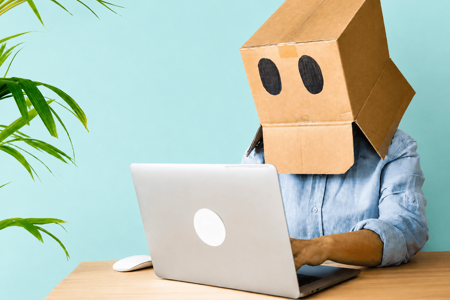 Une personne portant un masque en carton avec deux grands yeux noirs travaille sur un ordinateur portable sur un bureau en bois devant un mur bleu clair.
